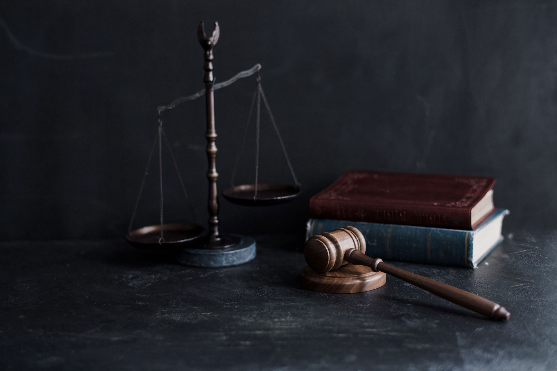 A wooden judges gavel rests beside a stack of two books and a set of balance scales on a dark, textured surface with a dark background.