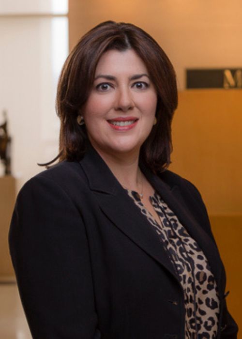 Sila Gonzalez Puerto Rico A woman with shoulder-length dark hair, wearing a black blazer over a leopard print blouse, smiles at the camera in an indoor setting with a blurred background.