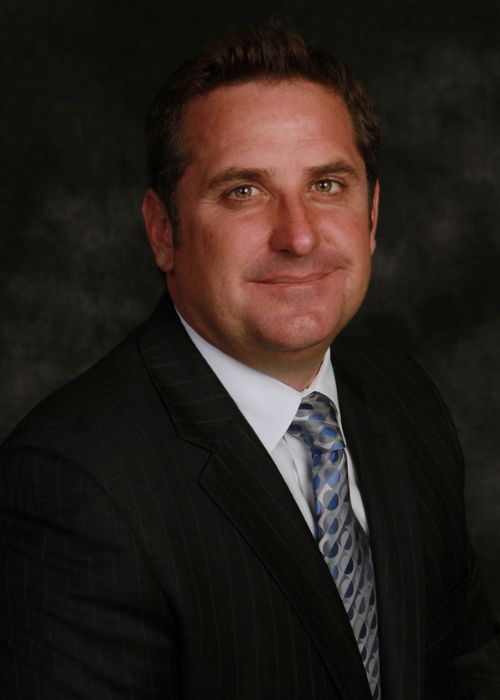 Todd Hill Minnesota A man in a dark pinstripe suit, white shirt, and patterned blue tie is smiling slightly against a dark, mottled background. He has short brown hair and is facing the camera.