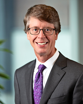A smiling man with short brown hair and glasses wearing a dark suit, white shirt, and purple patterned tie, standing indoors with a blurred office background.
