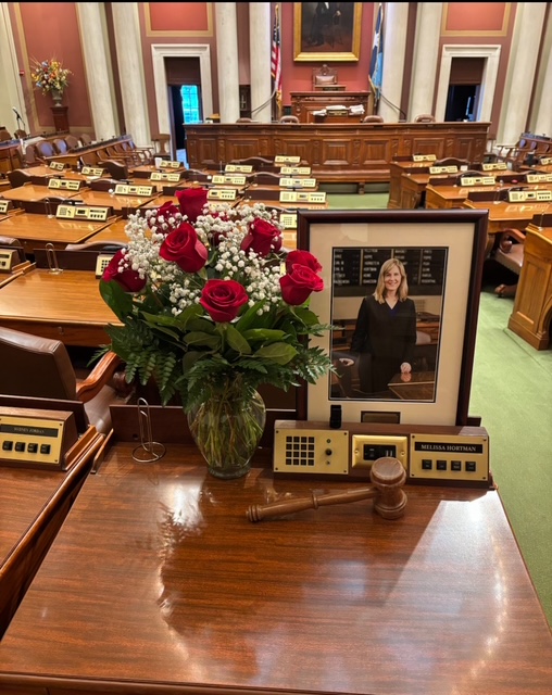 A wooden desk in a legislative chamber holds a framed photo of a woman, a bouquet of red roses with baby’s breath in a vase, a gavel, and a nameplate reading “Melissa Horman.” Rows of desks fill the background.