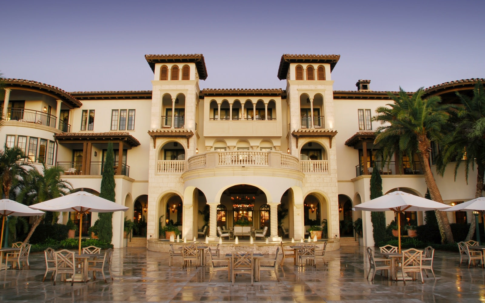 A grand Mediterranean-style building with arches, balconies, and tower-like structures, surrounded by palm trees. Outdoor dining tables and chairs sit on a stone patio in the foreground under large white umbrellas.
