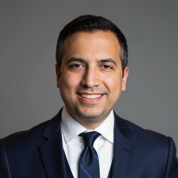 A man in a dark suit, white shirt, and dark tie is smiling at the camera against a plain gray background.
