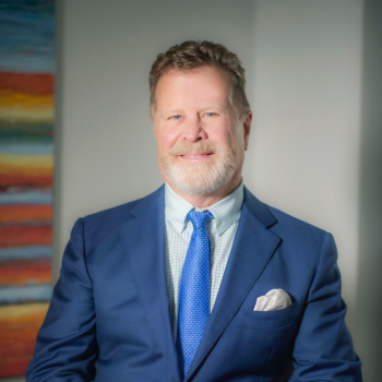 A man with light hair and a beard, wearing a blue suit, blue tie, and checkered shirt, smiles while standing indoors. A colorful abstract painting is visible in the background.