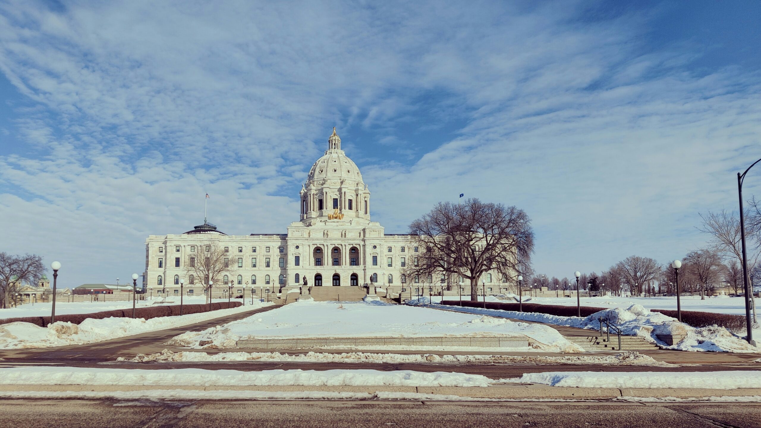 The image shows a large, domed government building in winter, surrounded by snow-covered ground, leafless trees, and a bright blue sky with scattered clouds.