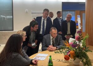 A group of people, including a man in a suit seated at a table with papers and a flower arrangement, look at a computer screen together in a modern indoor setting. Several others stand and watch, smiling.