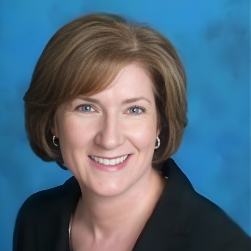 A woman with short light brown hair, wearing a black blazer and small hoop earrings, smiles at the camera against a blue background.