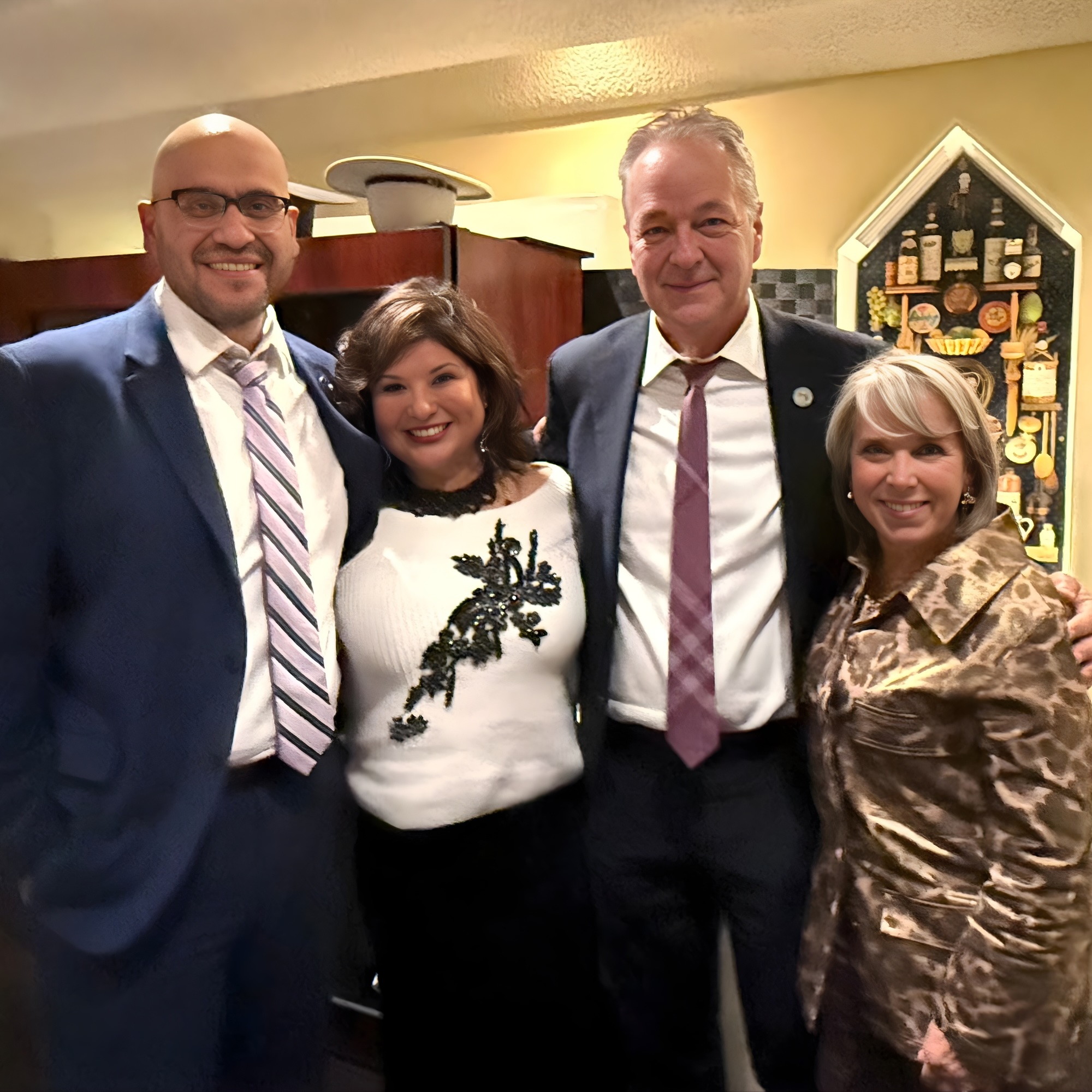 Four adults dressed in formal attire stand close together, smiling at the camera in a warmly lit indoor setting with kitchen decor visible in the background.