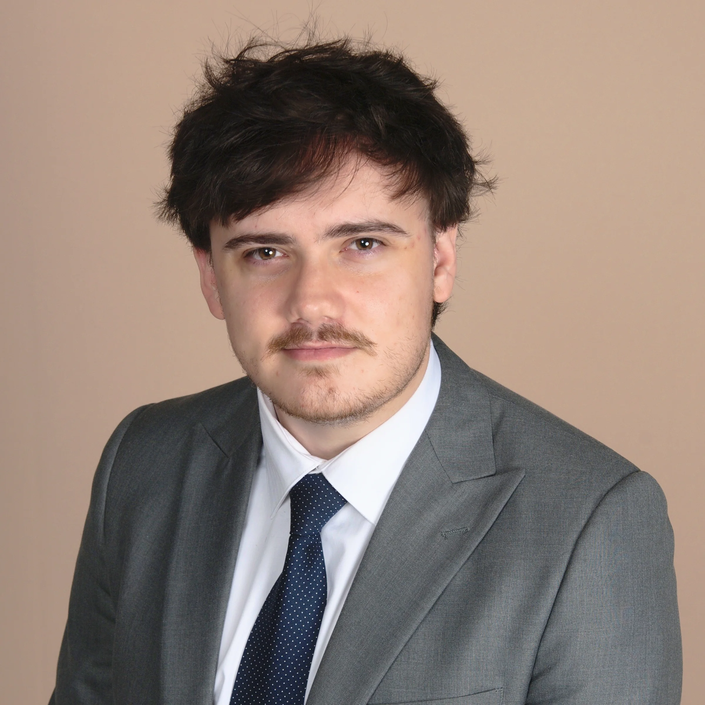 A young man with dark, tousled hair and a mustache wears a gray suit, white shirt, and dark tie. He is sitting and looking at the camera with a neutral expression against a beige background.