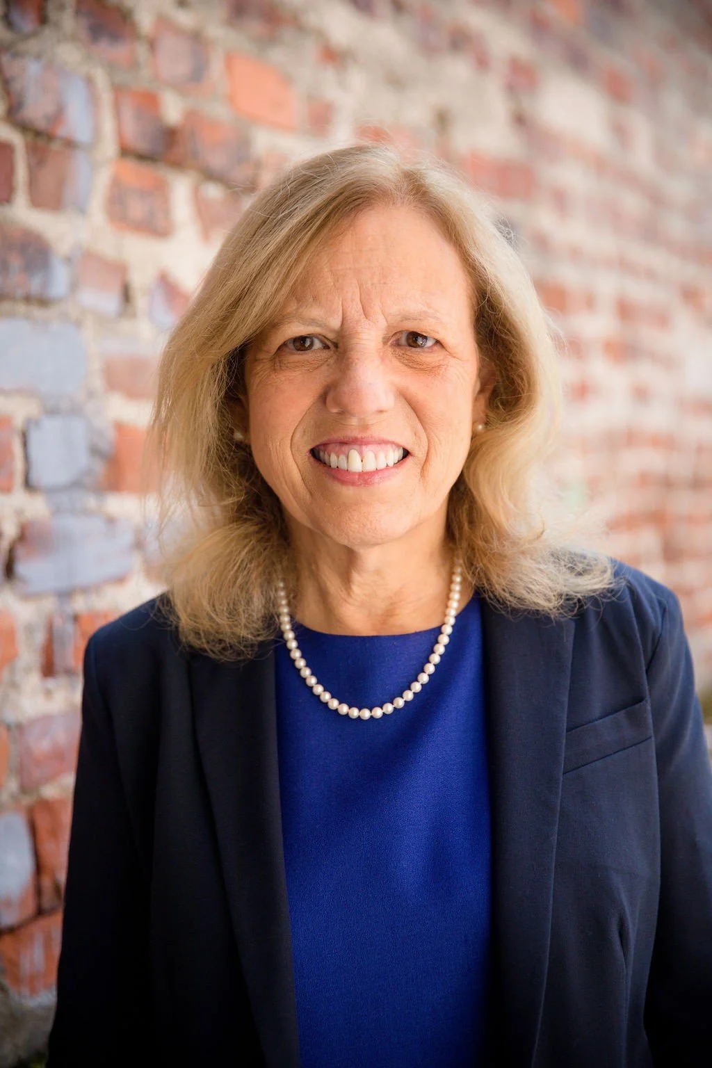 A woman with shoulder-length blonde hair, wearing a navy blazer, blue top, and a pearl necklace, smiles while standing in front of a red brick wall.