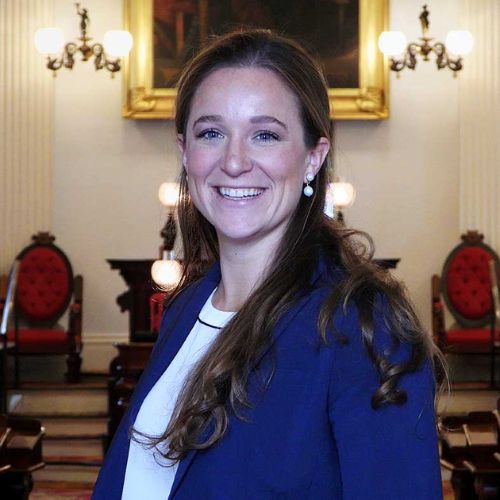 A woman with long brown hair, wearing a blue blazer and white top, smiles while standing in an elegant room with red chairs, chandeliers, and a large framed painting in the background.