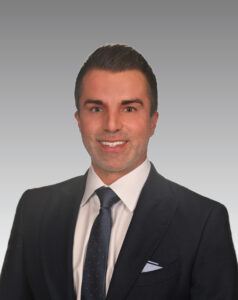 A man in a dark suit, white shirt, and dark tie is smiling at the camera against a gradient gray background.
