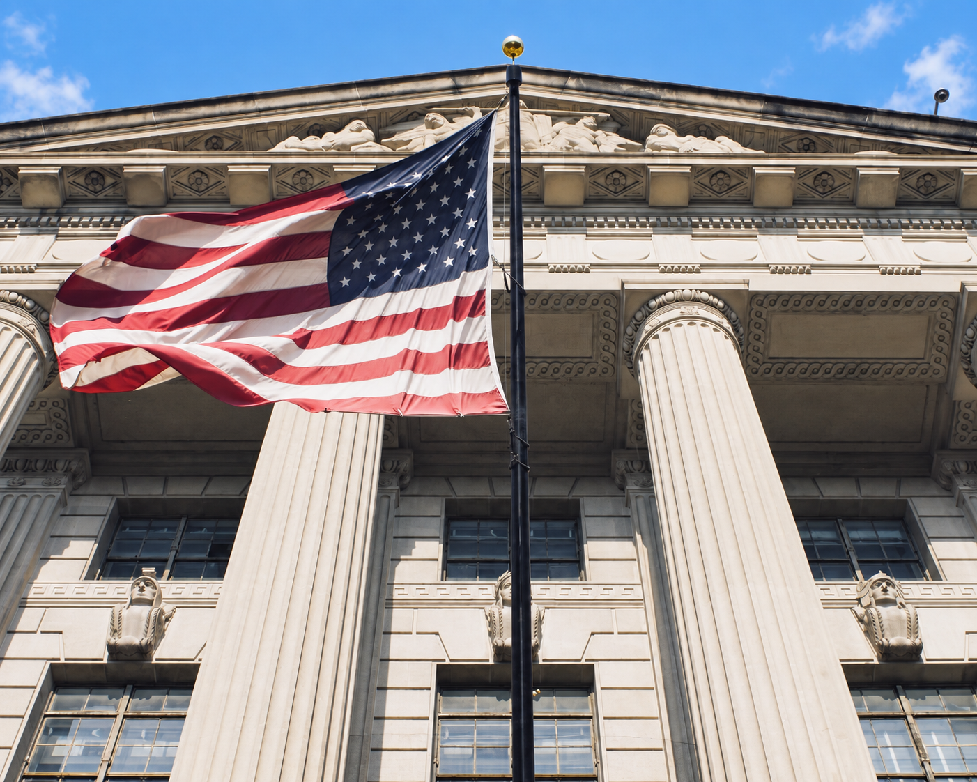 The U.S. flag waves in front of a neoclassical government building with tall columns and ornate stonework under a blue sky.
