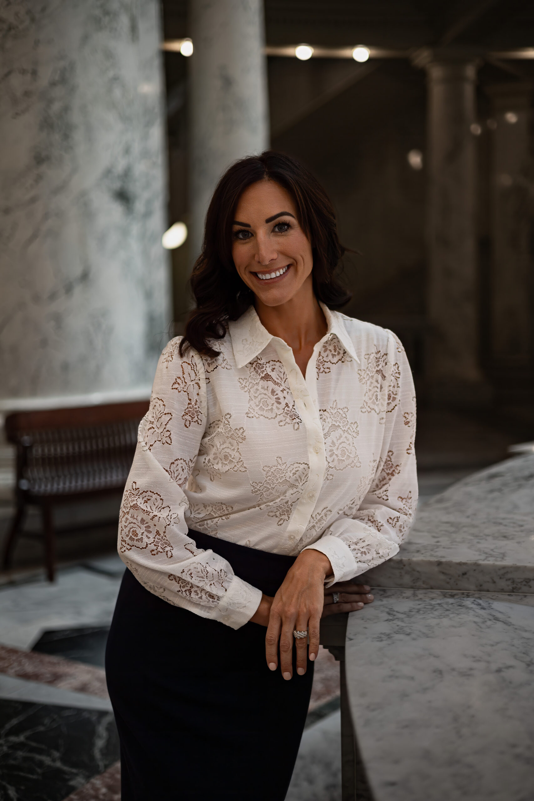 Carlie M. Foster - Idaho - New A woman with dark hair, wearing a white lace blouse and dark skirt, stands smiling and leaning on a marble counter in an elegant, marble-columned interior space.