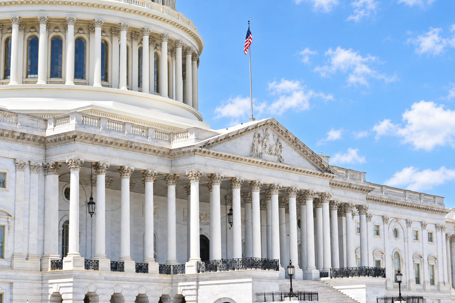 The U.S. Capitol building with white columns, a dome, and an American flag flying above, set against a blue sky with scattered clouds.