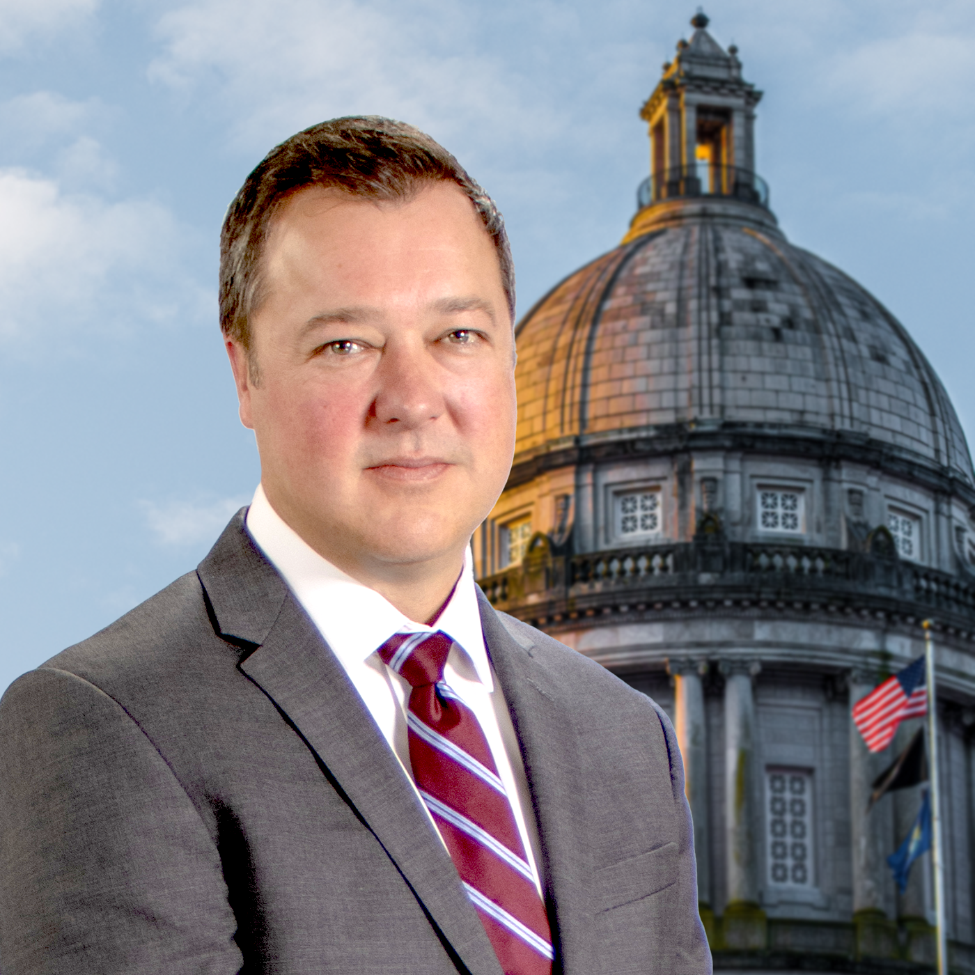 Jason Bentley A man in a gray suit and red striped tie stands in front of a domed government building with American flags, under a partly cloudy sky.