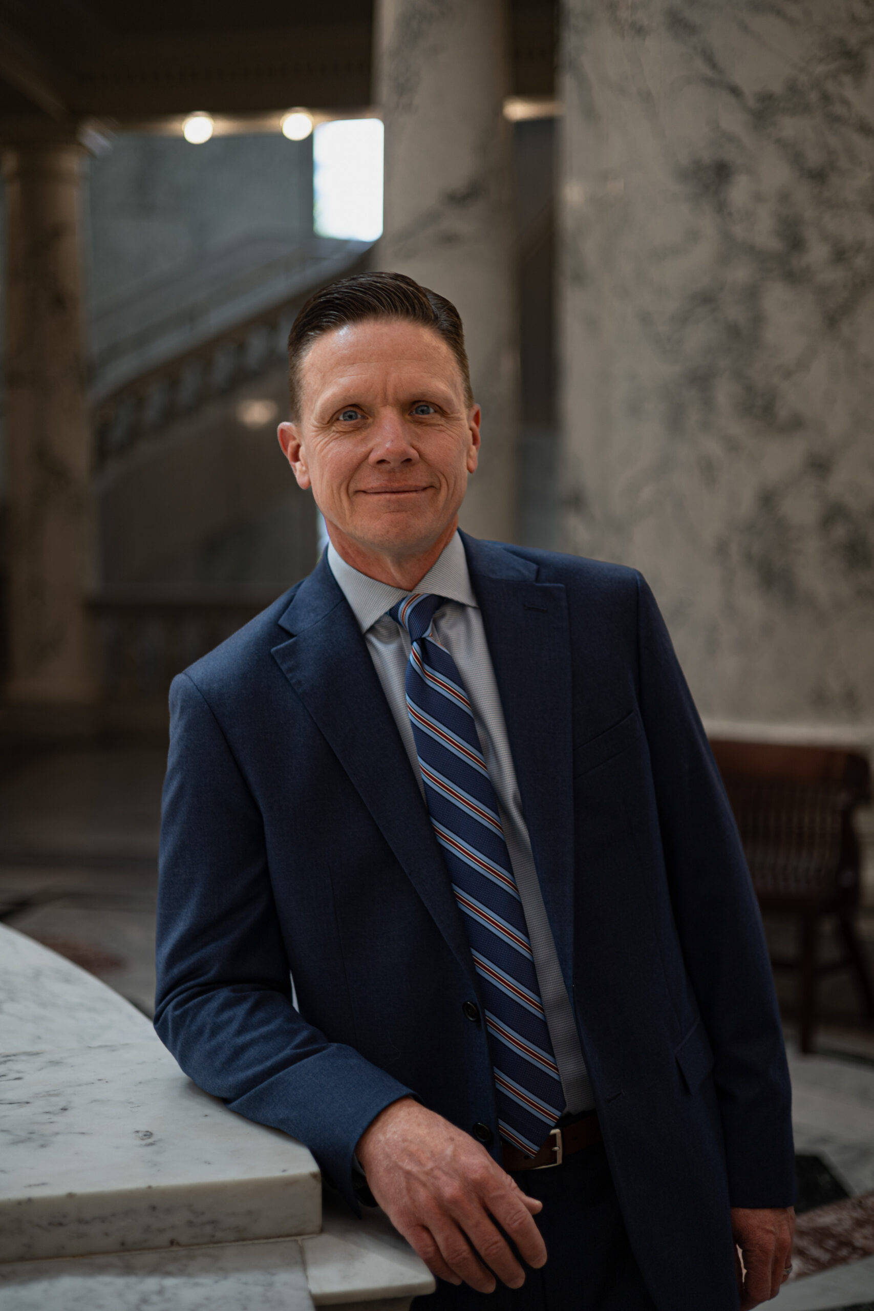 Jason Kreizenbeck - Idaho - New A man in a blue suit and striped tie leans on a marble surface in an elegant, marble-columned interior, smiling confidently at the camera.