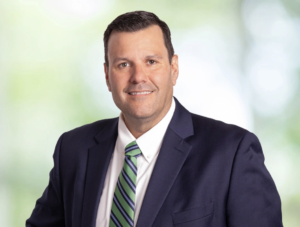 Man in a dark suit, white shirt, and green striped tie, smiling at the camera with a blurred light green and white background.