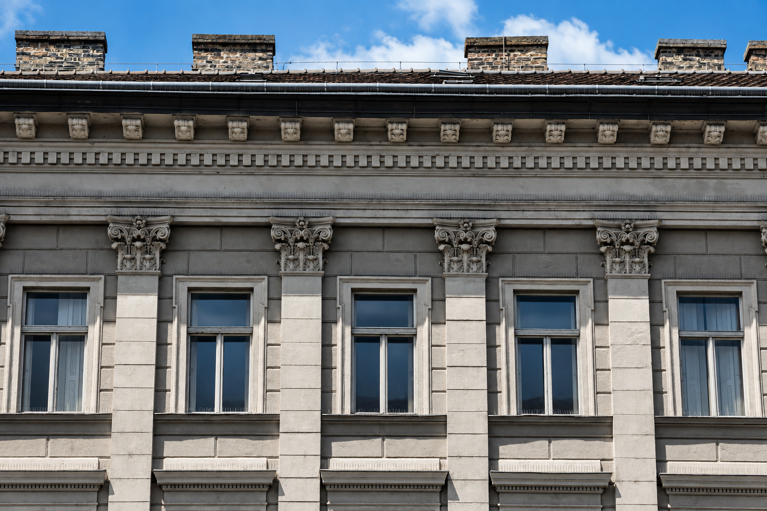 The image shows the upper facade of an old building with decorative columns, four windows, ornate architectural details, and a row of brick chimneys against a blue sky with some clouds.