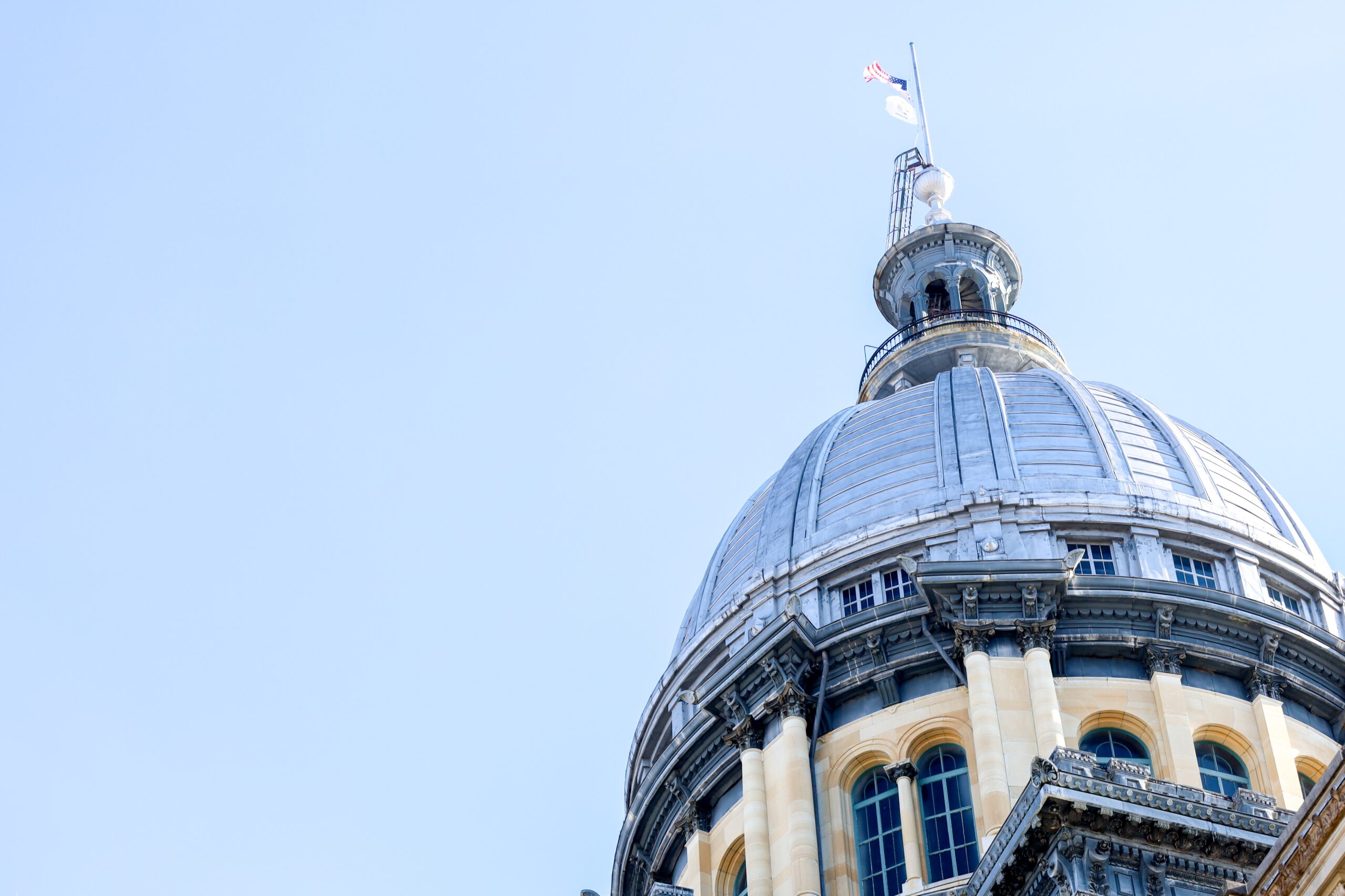 The top portion of a domed government building with detailed architecture and a flag at its peak, set against a clear blue sky.